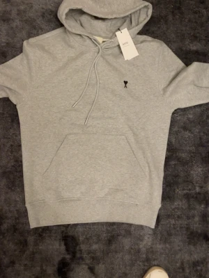 Grå hoodie från Ami Paris - Snygg ljusgrå hoodie från Ami Paris med klassisk huva och dragsnören. Har en känguruficka framtill och en liten svart Ami-logga broderad på bröstet. Perfekt för en chill och stilren look.