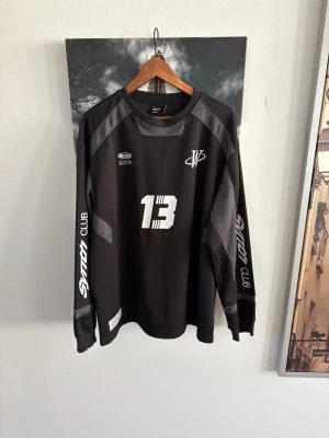 Svart långärmad tröja Synth Club - Svart långärmad sporttröja från Morton med tryckta vita detaljer, nummer 13 på bröstet och 'Synth Club' på ärmarna. Tröjan har grå paneler på axlar och ärmar samt ribbade muddar. Perfekt för dig som gillar sportig och unik stil.