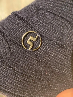 Mörkblå kabelstickad tröja J.Lindberg - Kabelstickad mörkblå tröja från J.Lindberg med rund halsringning och ribbade muddar. Tröjan har ett diskret metallmärke nertill och är perfekt för lager-på-lager. Klassisk och stilren design som funkar till många outfits.