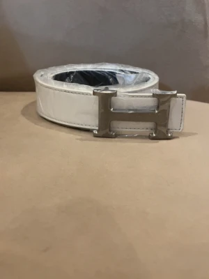 Vitt skärp med H spänne från H’er’m”e’s - Snyggt vitt skärp från H’er’m”e’s med  H spänne. Skärpet har en stilren, klassisk design som passar till många olika outfits.