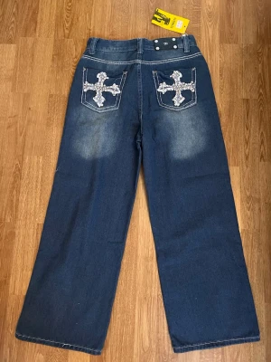 Blåa wide leg jeans med broderi - Säljer ett par blåa wide leg jeans från Southpole med coola broderade kors på bakfickorna. Jeansen har slitningar framtill och en avslappnad passform. Perfekta för dig som gillar streetwear och vill sticka ut med detaljer.