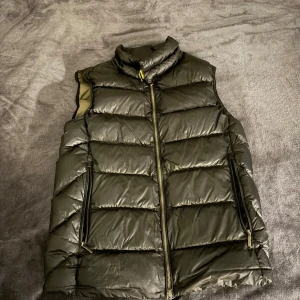 Svart dunväst Parajumpers - Stilren svart dunväst från Parajumpers med quiltad design och hög krage. Västen har dragkedja framtill, två snedställda fickor med dragkedja och insida i kontrasterande beige. Perfekt för lager-på-lager och streetstyle.