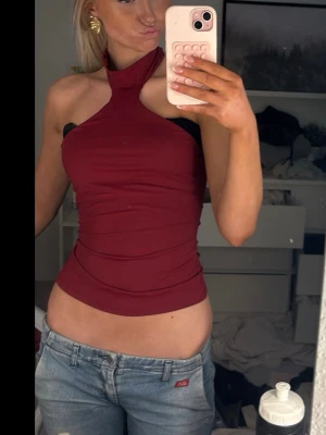 Vinröd halterneck topp - Säljer en vinröd halterneck topp med hög hals och ärmlös design. Toppen är tight och har en snygg, modern passform som framhäver axlarna. Perfekt att styla med jeans eller kjol för en trendig look. Assnygg topp men kommer tyvär inte till användning hos mig🥹 