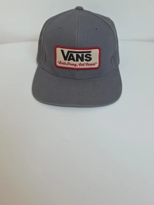 Grå keps från Vans med patch - Snygg grå keps från Vans med rak skärm och broderad patch framtill i vitt och rött med texten 'Built Strong, Cali Casual'. Klassisk modell i bomullsmaterial som passar perfekt till streetwear-stilen.