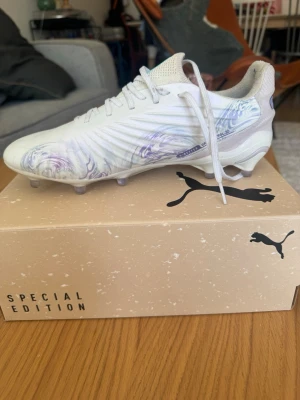 Puma Special Edition fotbollsskor - Säljer ett par vita Puma Special Edition fotbollsskor med unika lila och blå swirl-detaljer på sidan. Skorna har klassisk snörning, låg profil och är tillverkade i syntetmaterial för bästa känsla på planen. Perfekta för dig som vill sticka ut på matchen.