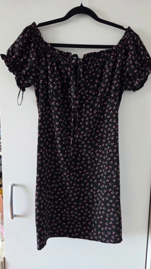 Svart blommig kortklänning - Supersöt svart kortklänning med små blommor i rött och vitt. Klänningen har puffärmar, snörning framtill och kan bäras offshoulder. Perfekt för sommarens alla tillfällen och ger en romantisk vibe. Hittar inte vilken storlek den är men troligtvis s/m