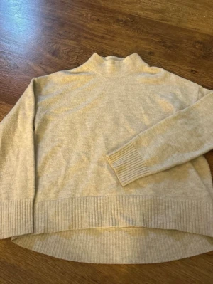 Beige stickad tröja från Kappahl - Mysig beige stickad tröja från Kappahl med hög krage och långa ärmar. Tröjan har ribbade muddar vid ärmslut och nederkant samt en något längre bakdel. Perfekt för lager på lager och enkel att matcha till olika outfits. Knottrig annars bra skick