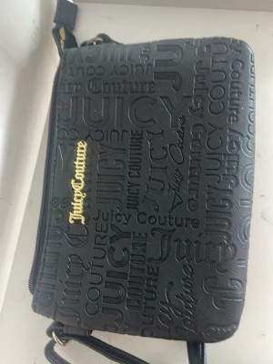 Svart Juicy Couture handväska - Snygg svart handväska från Juicy Couture i konstläder med präglad logotyp över hela väskan och guldfärgad Juicy Couture-text på framsidan. Väskan har justerbar axelrem och dragkedja upptill. Perfekt för dig som gillar streetstyle och vill ha en trendig accessoar.