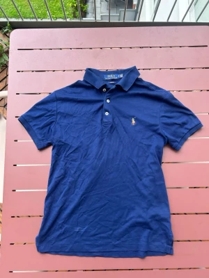 Marinblå piké från Polo Ralph Lauren - Klassisk marinblå pikétröja från Polo Ralph Lauren med korta ärmar och den ikoniska broderade loggan på bröstet. Tillverkad i mjuk bomull och har krage med knappar framtill. Perfekt för en stilren och avslappnad look.