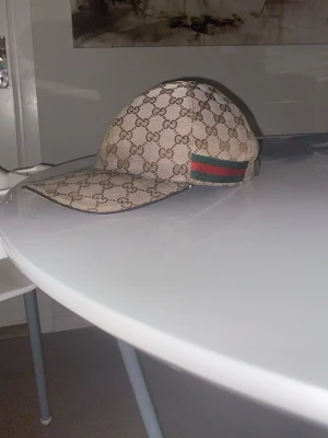 Beige keps från Gucci med GG-mönster - Säljer en beige keps från Gucci med det klassiska GG-monogrammet över hela kepsen. På sidan finns en grön och röd rand som signaturdetalj. Kepsen har böjd skärm och justerbar rem bak. Materialet är canvasliknande tyg.