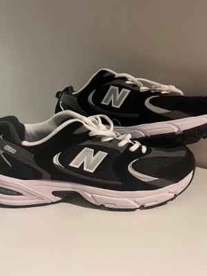 New Balance 530 svarta sneakers - Svarta New Balance 530 sneakers med vita och grå detaljer. Skorna har en klassisk låg siluett, meshpaneler för ventilation och vita snören. Mellansulan är vit med svart yttersula och märkets ikoniska N-logga på sidan.