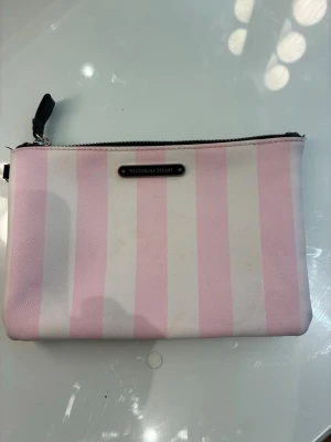 Rosa och vit randig  necessär från Victoria Secret - Snygg  necessär från Victoria Secret med klassiska rosa och vita ränder. Necessären har en svart dragkedja och en metallbricka med loggan framtill. Praktiskt material som går att torka av/tvätta!  Storlek: 18cm*12 cm. Necessären har några mindre skönhetsfläckar av från smink på utsidan(se bilder), Insidan är svart och i perfekt skick, likaså dragkedjan. Priset är förhandlingsbart!😊