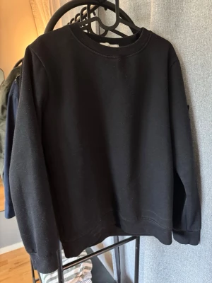 Svart sweatshirt från Stone Island - Säljer en svart sweatshirt från Stone Island med klassisk rund hals och ribbade muddar. Tröjan har den ikoniska Stone Island-loggan på vänster ärm. Perfekt för en stilren och avslappnad look.