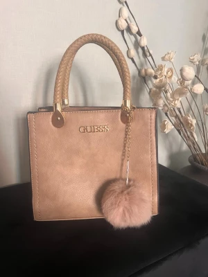 Beige handväska från Guess med fluffig detalj - Snygg beige handväska från Guess med guldiga detaljer och flätade handtag. Väskan har en fluffig beige nyckelring, dragkedjeficka på baksidan och stilren fyrkantig form. Perfekt för dig som vill ha en trendig och praktisk accessoar.De är inte riktig guess men ser exakt ut som en riktig 