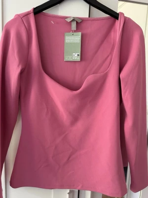 Rosa långärmad topp från H&M - Trendig rosa långärmad topp från H&M med djup, rundad urringning. Tillverkad i mjuk bomullsmix som känns skön mot huden. Perfekt att styla med jeans eller kjol för en snygg och enkel look.