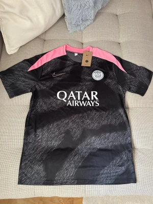 Svart PSG matchtröja från Nike - Snygg svart PSG matchtröja från Nike med rosa detaljer på axlarna och vitt Qatar Airways-tryck på bröstet. Tröjan har klubbmärke och Nike-logga framtill, samt ett mönster i grått över hela tyget. Tillverkad i lätt och ventilerande material, perfekt för fotboll eller träning.