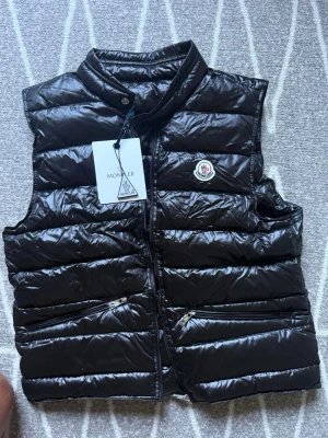 Moncler väst - Säljer en helt ny Snygg svart dunväst från Moncler med glansig finish och klassisk logotyp på bröstet. Västen har hög krage, dragkedja framtill och två fickor med dragkedja. Dustbag och ettikter tillkommer såklart. (pris kan diskuteras)