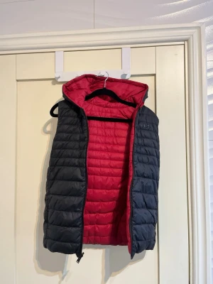 Svart och röd hugo boss väst - Snygg dunväst i svart med röd insida och vadderad design. Västen har en stor huva och dragkedja framtill. Perfekt för lager-på-lager och håller dig varm under kyliga dagar. Materialet är lätt och quiltat för extra komfort. Två sidig väst