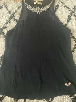 Svart linnetopp med strass från Hollister - Snygg svart linnetopp från Hollister med glittrande strassdetaljer runt halsringningen. Toppen är ärmlös och har en liten Hollister-logga broderad nertill. Perfekt för dig som vill ha något stilrent men ändå extra.