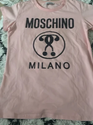 Rosa Moschino Milano T-shirt - Snygg rosa T-shirt från Moschino med stort svart Moschino Milano-tryck framtill. Klassisk rund halsringning och korta ärmar. Perfekt för dig som vill ha en trendig och bekväm tee med exklusiv känsla. Tillverkad i mjuk bomull. HELT OANVÄND 