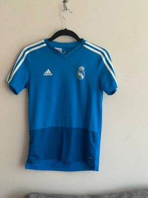 Blå Real Madrid träningströja Adidas - Snygg blå träningströja från Adidas med Real Madrid-logga på bröstet. Tröjan har vita ränder på axlarna, korta ärmar och V-ringning. Perfekt för fotbollsträning eller gymmet. Tillverkad i lätt och snabbtorkande material. Tröjan är i storlek 164 Barn