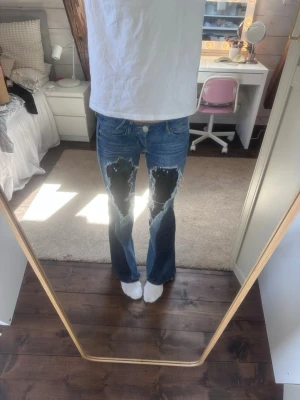 Bootcut jeans med paljetter - Säljer ett par blå bootcut jeans med stora slitningar och hål på framsidan av benen. Jeansen har klassisk femficksmodell och råa kanter vid bensluten. Perfekta för dig som gillar en edgy och trendig stil.