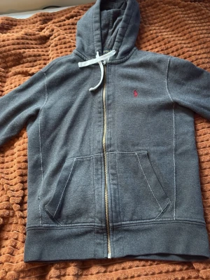 Mörkgrå zip hoodie med huva - Mörkgrå hoodie med dragkedja och huva, vita dragsnören och två stora fickor framtill. Liten röd logga broderad på bröstet. Tillverkad i mjukt sweatshirtmaterial, perfekt för chill dagar eller när du vill ha en enkel och stilren look.