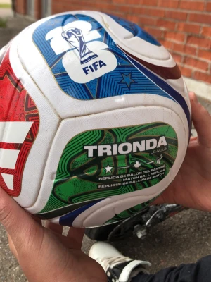 Adidas Trionda fotboll FIFA design - Säljer en Adidas Trionda League fotboll med FIFA-design. Bollen har en vit bas med färgglada detaljer i rött, blått, grönt och brunt samt tydlig Adidas-logga. Tillverkad i slitstarkt syntetmaterial och passar perfekt till träning eller match.
