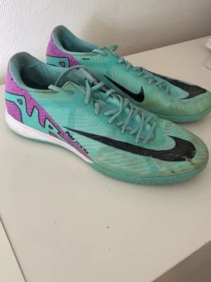 Nike inomhusskor - Ett par fotbollsskor från Nike i turkos med lila och svarta detaljer. Skorna har snörning framtill och en sula anpassad för inomhusbruk eller hårda underlag. Hyfsat skick funkar felfritt inget fel på dom. Lite slitage men annars inget.