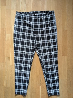 Svarta leggings med rutigt mönster - Säljer ett par svarta leggings med vitt rutigt mönster. De har hög midja och är stretchiga för en bekväm passform. Materialet känns mjukt och följsamt, perfekt för dig som gillar en trendig och avslappnad stil.