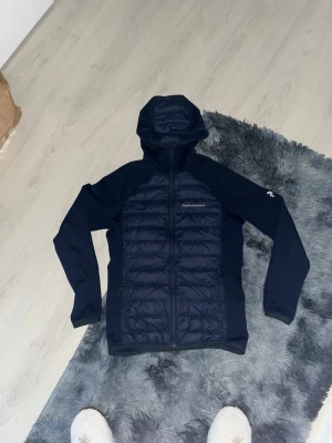 Peak performance mörkblå vindjacka med huva  - Snygg mörkblå vindjacka från Peak Performance med quiltad front och rygg, samt släta ärmar och sidor. Jackan har huva, dragkedja framtill och två sidofickor med dragkedja. Perfekt för blåsiga dagar och outdoor-aktiviteter.