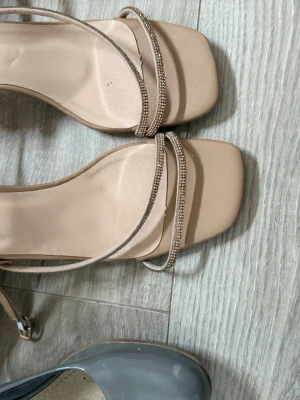 Beige pumps med strassdetaljer - Snygga beige pumps med fyrkantig tå och tunna remmar dekorerade med strass. Skorna har en elegant och minimalistisk design som passar perfekt till olika outfits. Tillverkade i ett slätt material med justerbar rem runt ankeln.
