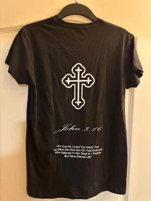 Svart t-shirt med kors och text - Svart t-shirt från NLA med vitt kors-tryck på bröstet och större kors på ryggen. Under korset på ryggen står bibelcitatet John 3:16 i vit text. Klassisk rund hals och korta ärmar. Tillverkad i mjuk bomull för skön känsla.