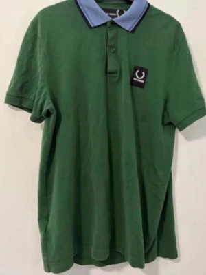 Fred Perry x Raf Simons Polo Shirt – BRAND NEW - Snygg grön pikétröja från Raf Simons med blå krage och svart patch med logga på bröstet. Klassisk passform med korta ärmar och knappar vid halsen. Perfekt för en clean och stilren look.