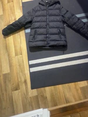 Svart pufferjacka med huva - Snygg svart pufferjacka med huva och dragkedja framtill. Jackan har två sidofickor med dragkedja och quiltad design som ger en klassisk look. Perfekt för kalla dagar och enkel att matcha med olika outfits.