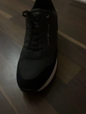 Svarta sneakers från Tommy Hilfiger - Snygga svarta sneakers från Tommy Hilfiger med klassisk design. Skorna har snörning och en vit sula som kontrast. Materialmix med inslag av mocka och syntet ger en modern look. Perfekta för dig som vill ha stilrena och bekväma sneakers.