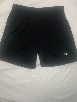Svarta tränings shorts fråm champion - Svarta shorts från Champion med elastisk midja och snörning. Tillverkade i ett lätt och bekvämt polyestermaterial, perfekta för träning eller chill. Klassisk Champion-logga broderad på ena benet. Snygg och enkel design som funkar till det mesta.