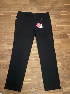 Svarta raka kostym/träningas byxor från Puma - Snygga svarta jeans från Puma med rak passform och klassisk femficksdesign. Jeansen har bälteshällor, knappgylf och är tillverkade i ett mjukt och slitstarkt material. Perfekta för en clean och stilren look.