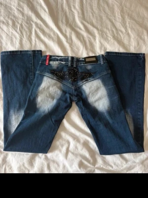 Blå jeans med broderad bakficka - Säljer ett par blå jeans med coolt broderi och nitar på bakfickan i form av en vinge. Jeansen har slitna detaljer och ljusa partier på baksidan. Klassisk femficksmodell med raka ben och normal midja. Perfekta för dig som vill sticka ut lite extra. Midjemått: 37,5cm Innerben: 81cm Försmå så kan ej visa på!