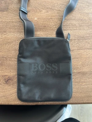 Hugo boss sidoväska - Snygg svart axelväska från Hugo Boss med stilren design och präglad logga på framsidan. Väskan har justerbar axelrem och dragkedja upptill. Fick den i present när jag var 10 typ använd kanske fem gånger skriv vid frågor och tveka inte !