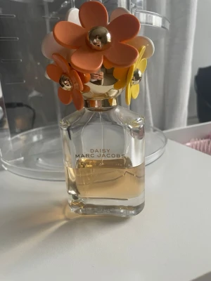 Daisy Eau So Fresh parfym Marc Jacobs - Daisy Eau So Fresh från Marc Jacobs är en parfym i en genomskinlig glasflaska med guldtext. Locket är dekorerat med stora plastblommor i orange, gult och vitt med gulddetaljer. Doften är fräsch och flaskan har en lekfull och färgglad design.