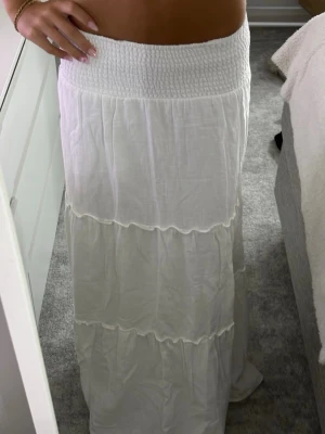 Vit långkjol med smock i midjan - Säljer en vit långkjol med smockad midja och volangdetaljer. Kjolen är luftig och har flera lager med lätt rynkade sömmar. passar mig som är 165 den är hyfsat för lång!