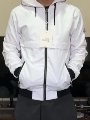 Vit vindjacka från Moncler med huva - Snygg vit vindjacka från Moncler med svart dragkedja och svarta detaljer vid fickor och midja. Jackan har huva med röda och blå ränder samt Moncler-logga på ärmen. Perfekt för blåsiga dagar och enkel att matcha med olika outfits.