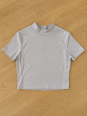 Beige ribbad croppad topp från SHEIN - Trendig croppad topp från SHEIN i beige färg med ribbad struktur. Toppen har korta ärmar och en högre krage som ger en clean och stilren look. Perfekt att matcha med höga jeans eller kjol för en enkel men snygg outfit.