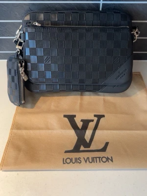 Svart axelväska Damier - Snygg svart axelväska i Damier-mönstrat läder. Väskan har silverfärgade detaljer, två dragkedjefack och en matchande liten plånbok. Perfekt för dig som gillar exklusiva accessoarer med stilren design.