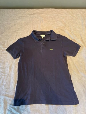 Mörkblå pikétröja från Lacoste - Klassisk mörkblå pikétröja från Lacoste med korta ärmar och den ikoniska gröna krokodilloggan på bröstet. Tröjan har krage och knappar vid halsen. Perfekt till en avslappnad men ändå stilren look.