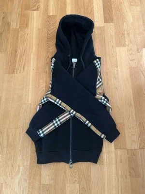 Svart Burberry hoodie med dragkedja helt ny  - Svart hoodie från Burberry med dragkedja framtill och klassiskt rutmönster längs ärmarna och sidorna. Huva med snörning och detaljer i metall. Perfekt för dig som vill ha en stilren och lyxig look med streetkänsla. Billigast på plick!! 