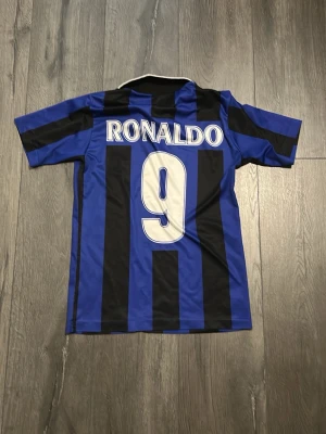 Inter Milan fotbollströja Ronaldo 9 - Klassisk Inter Milan fotbollströja i blått och svart med vertikala ränder, korta ärmar och krage. På framsidan finns Pirelli-loggan och klubbmärke, på baksidan står det Ronaldo och nummer 9 i vitt. Perfekt för dig som älskar fotboll och retrostil.