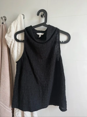 Svart ärmlös topp med hög krage - Säljer en svart ärmlös topp med hög krage och strukturerat tyg. Toppen har en lös passform och är tillverkad i ett lätt, något transparent material som ger en cool look. Perfekt att styla med jeans eller kjol för en trendig outfit.