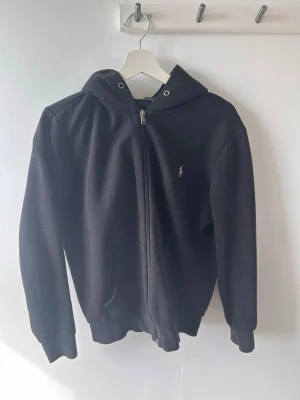 Svart hoodie från Ralph Lauren - Säljer en svart hoodie från Ralph Lauren med dragkedja framtill och klassisk huva. Tröjan har fickor på sidorna och den ikoniska lilla loggan broderad på bröstet. Perfekt för dig som gillar stilrena och bekväma plagg.